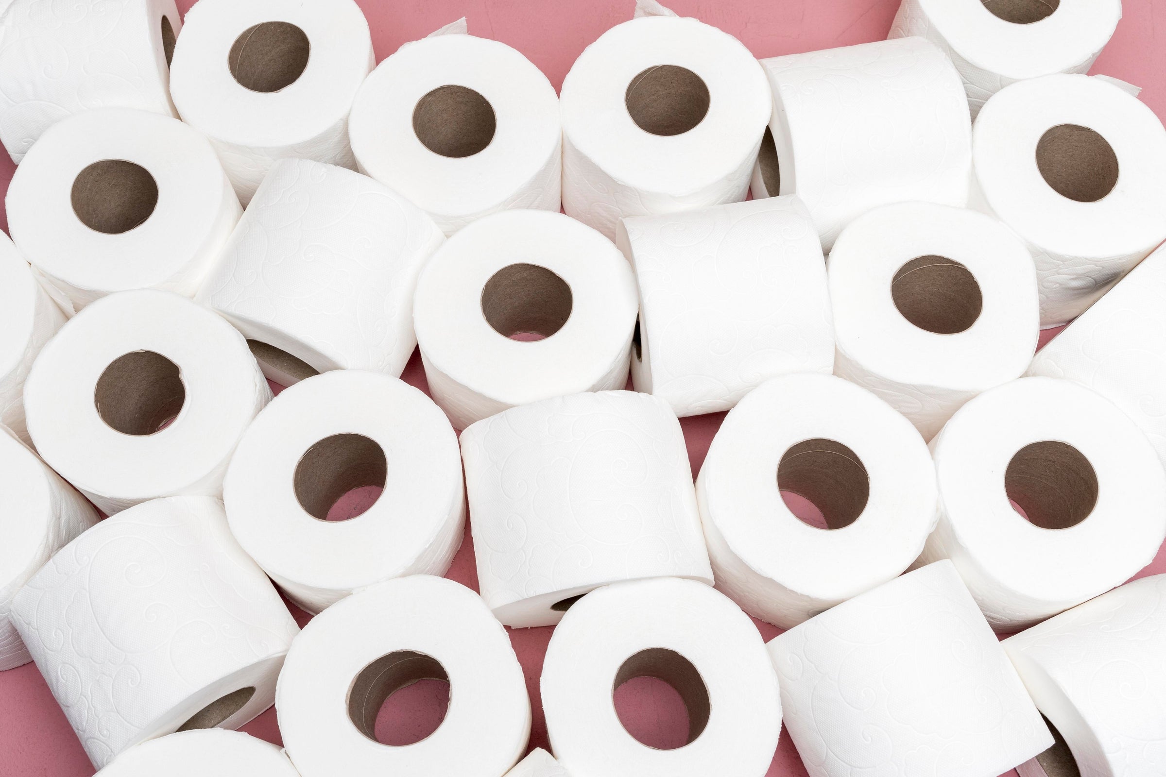 Un grand nombre de rouleaux de papier toilette blancs empilés pêle-mêle sur un fond rose, illustrant l'abondance et la propreté.